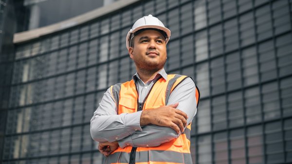 Safety manager wearing bright vest and hard hat standing with arms crossed in front of a building