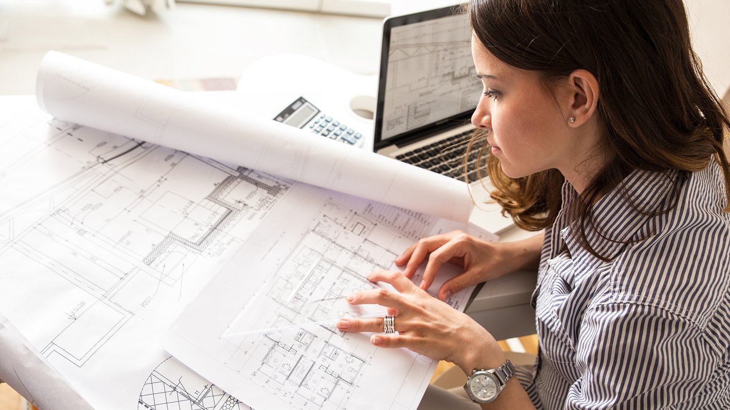 Female construction engineer reviewing architectural blueprints at a desk with a laptop.