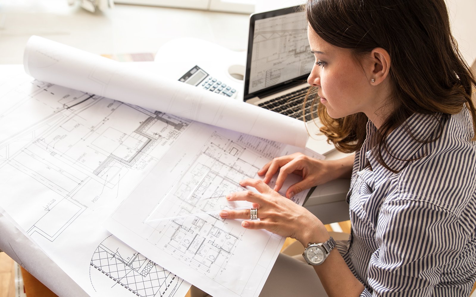 Female construction engineer reviewing architectural blueprints at a desk with a laptop.