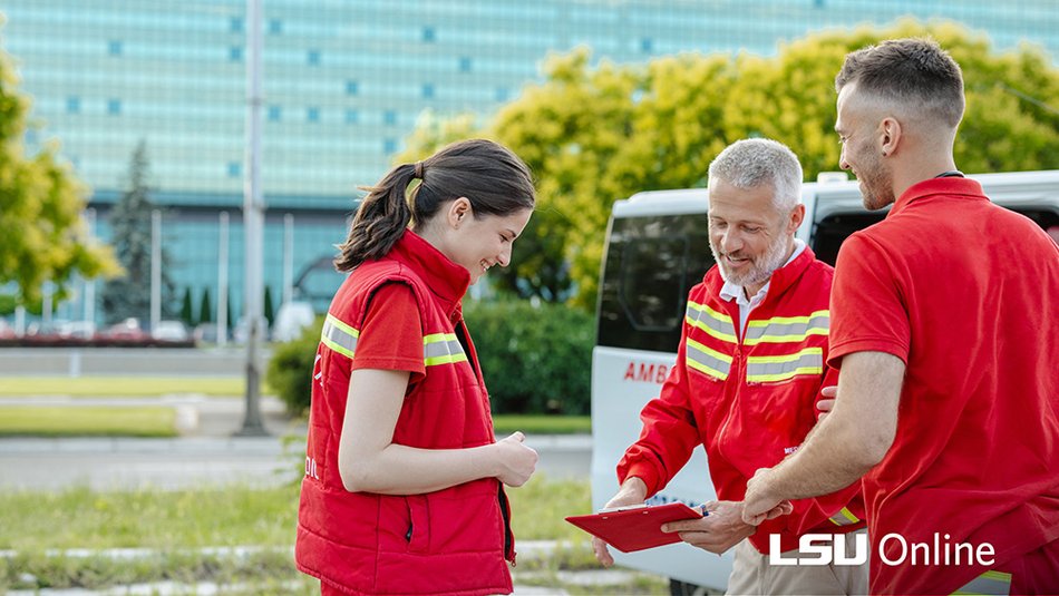 Three disaster response professionals in red emergency uniforms review a clipboard together outside near an ambulance, representing teamwork and coordination in a career in disaster preparedness and response.