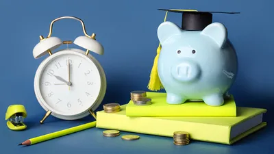Blue piggy bank with a graduate cap on sitting on yellow books next to an alarm clock