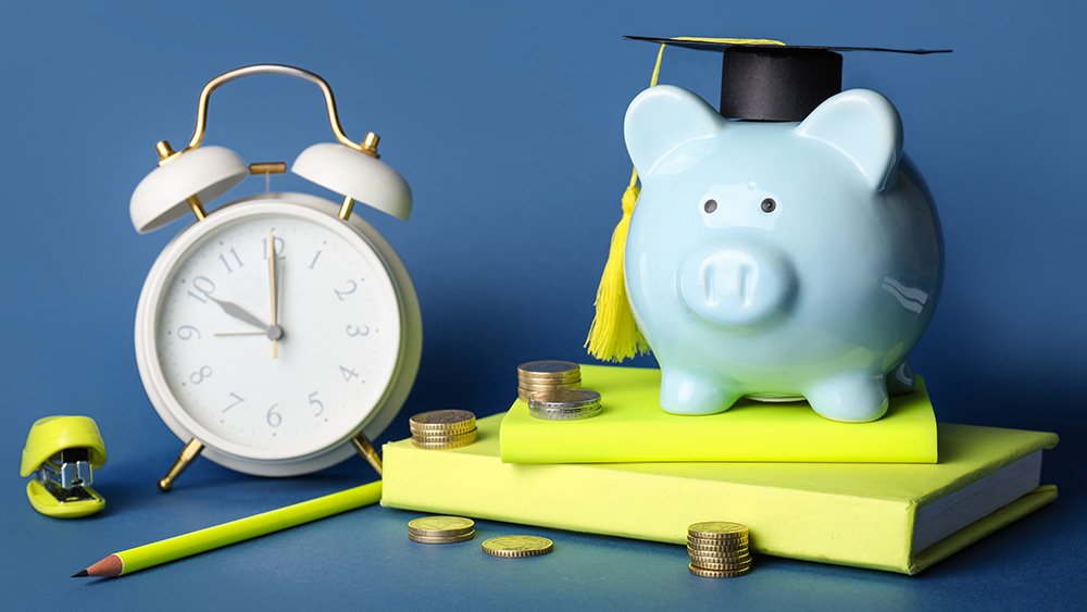 Blue piggy bank with a graduate cap on sitting on yellow books next to an alarm clock