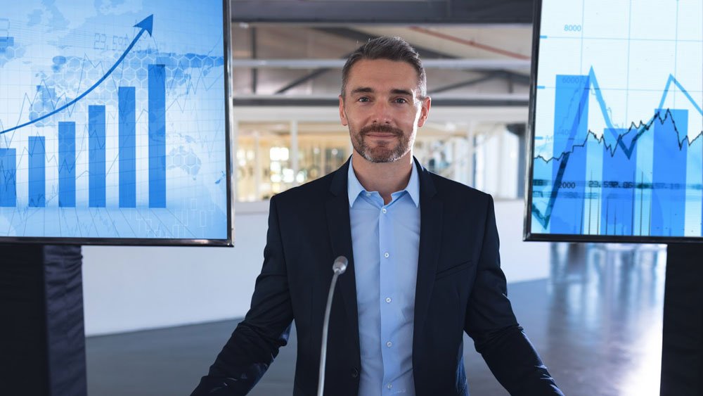 Man stands at podium in front of various charts and graphs.