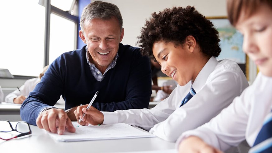 A teacher focuses on work with a student while at a desk.