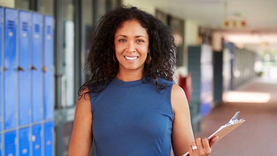 A woman with curly dark brown hair and wearing a sleeveless blue dress smiles at the camera while holding a clipboard.