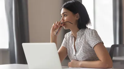 A woman with dark hair sits in front of a laptop computer, holding glasses.