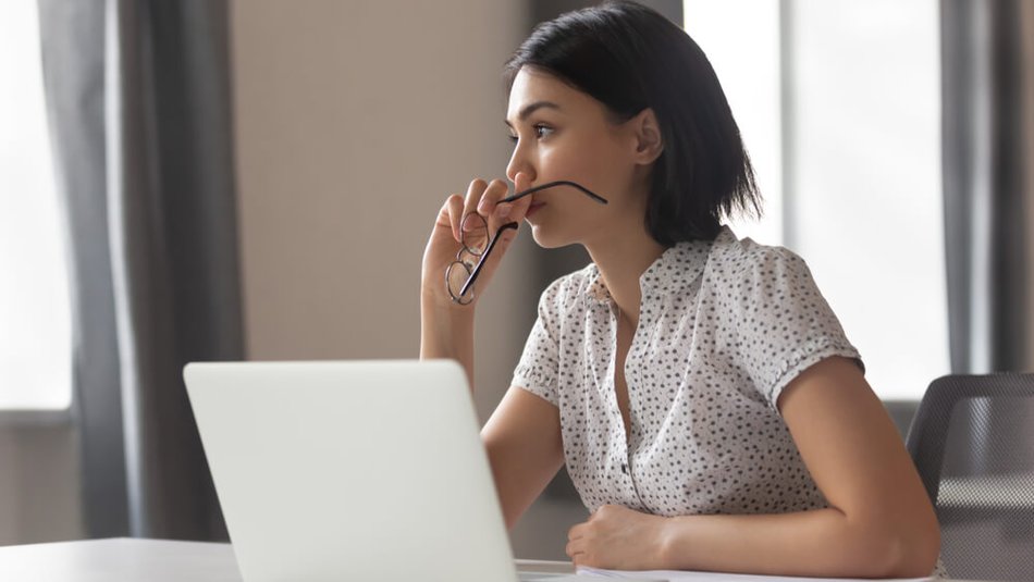 A woman with dark hair sits in front of a laptop computer, holding glasses.