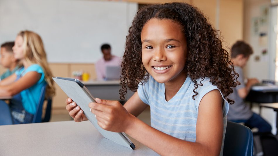 Smiling elementary school girl at classroom desk works with a tablet.