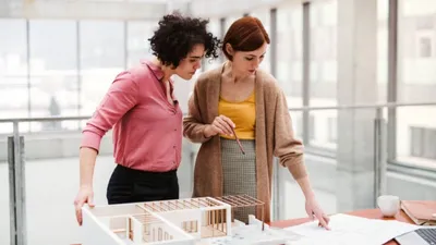Two women in construction management stand in an office next to a model of a building on a table, discussing a blueprint.