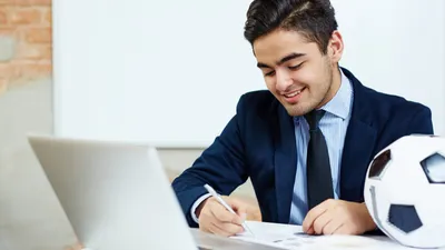 Man in a suit writing near a computer with a soccer ball on the desk