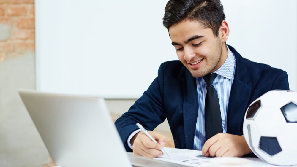 Man in a suit writing near a computer with a soccer ball on the desk